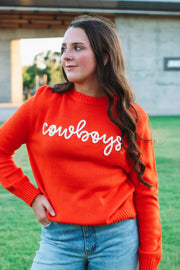 Deep orange hued sweater with 3D rope embroidery in white that says ‘cowboys’.