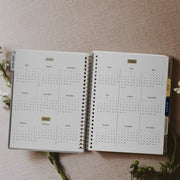 Open spiral-bound calendar with 2026 layout on a textured surface with flowers.