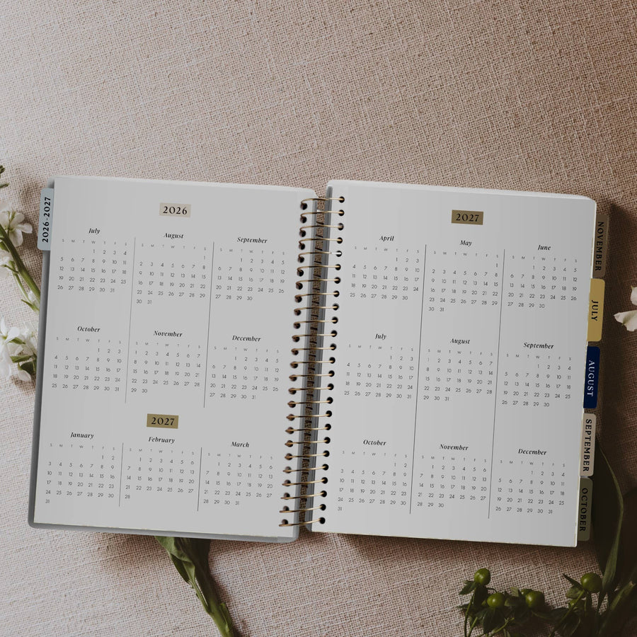 Open spiral-bound calendar with 2026 layout on a textured surface with flowers.