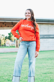 Deep orange hued sweater with 3D rope embroidery in white that says ‘cowboys’.