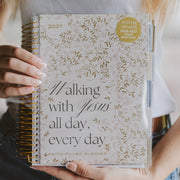 Planner with floral design and text 'Walking with Jesus all day, every day' held by a person.