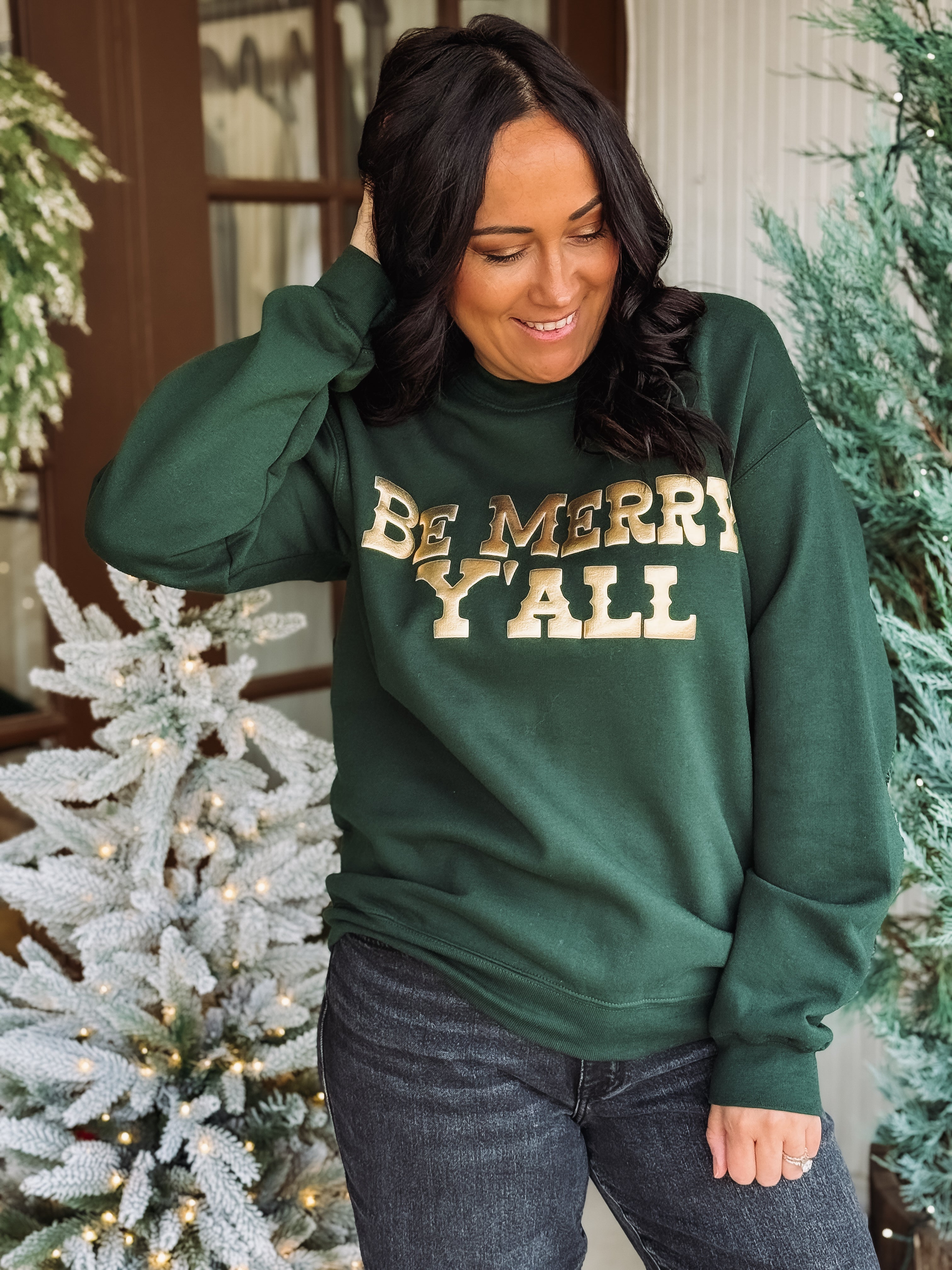 'Be Merry Y'all' printed in gold shiny lettering on a forest green crewneck.