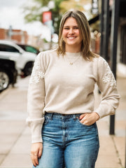 Woman wearing a beige sweater with white floral embroidered  sleeves.