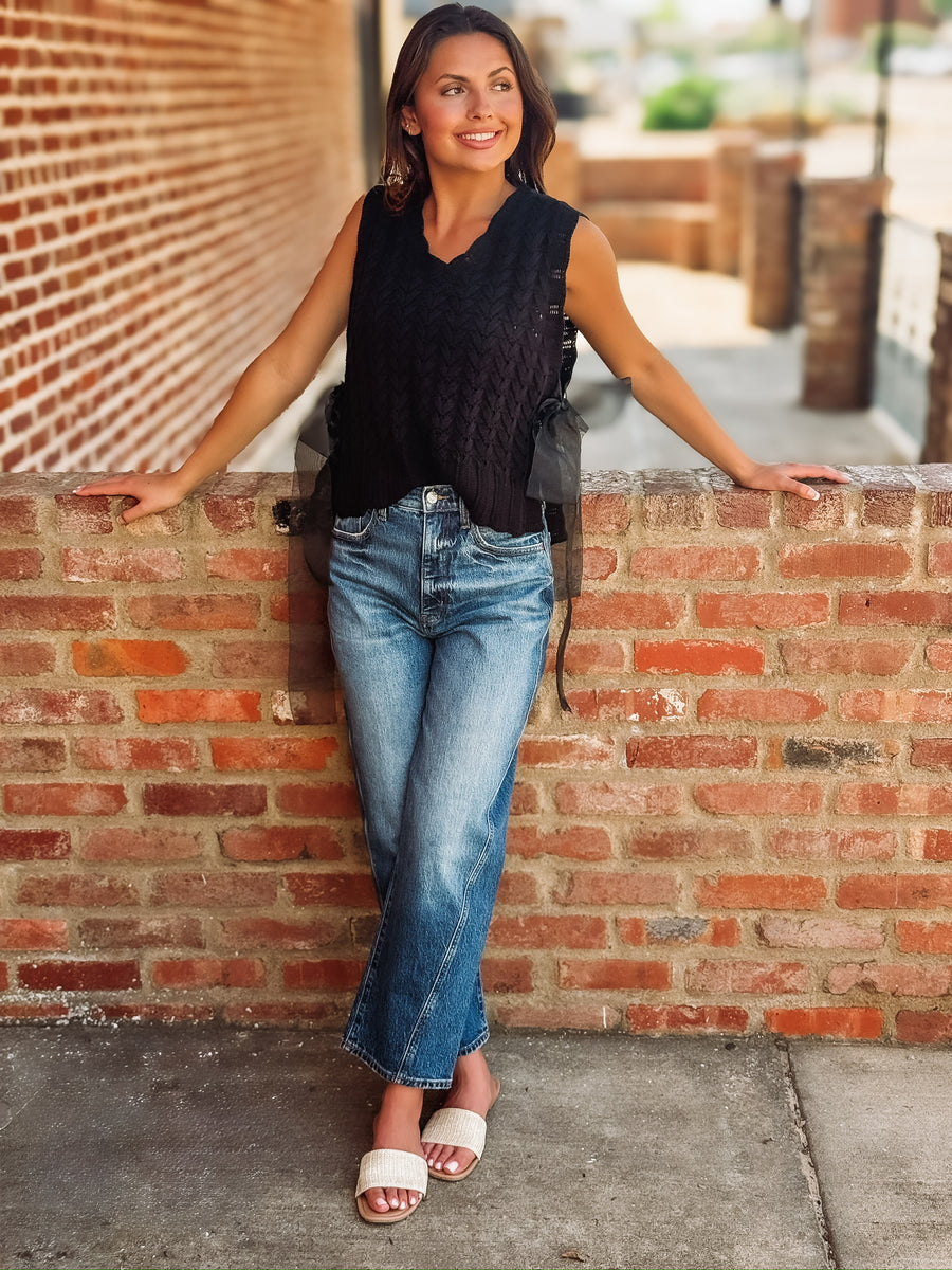 Black textured knit sweater vest with a v-neckline, ribbed trim, and open sides finished with adjustable sheer tulle tie details. Lightweight knit that can be worn alone or layered.