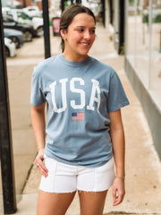 Woman wearing a blue 'USA' t-shirt with an American flag on a sidewalk.