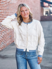 Woman wearing a white sweater with blue trim and blue jeans standing against a brick wall.