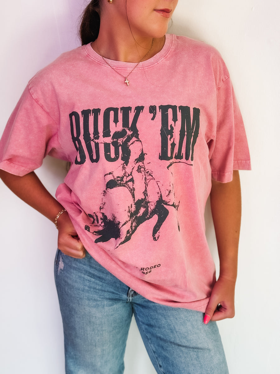 'Buck 'Em' in a western font with a bull rider graphic on an oversized washed pink shirt.