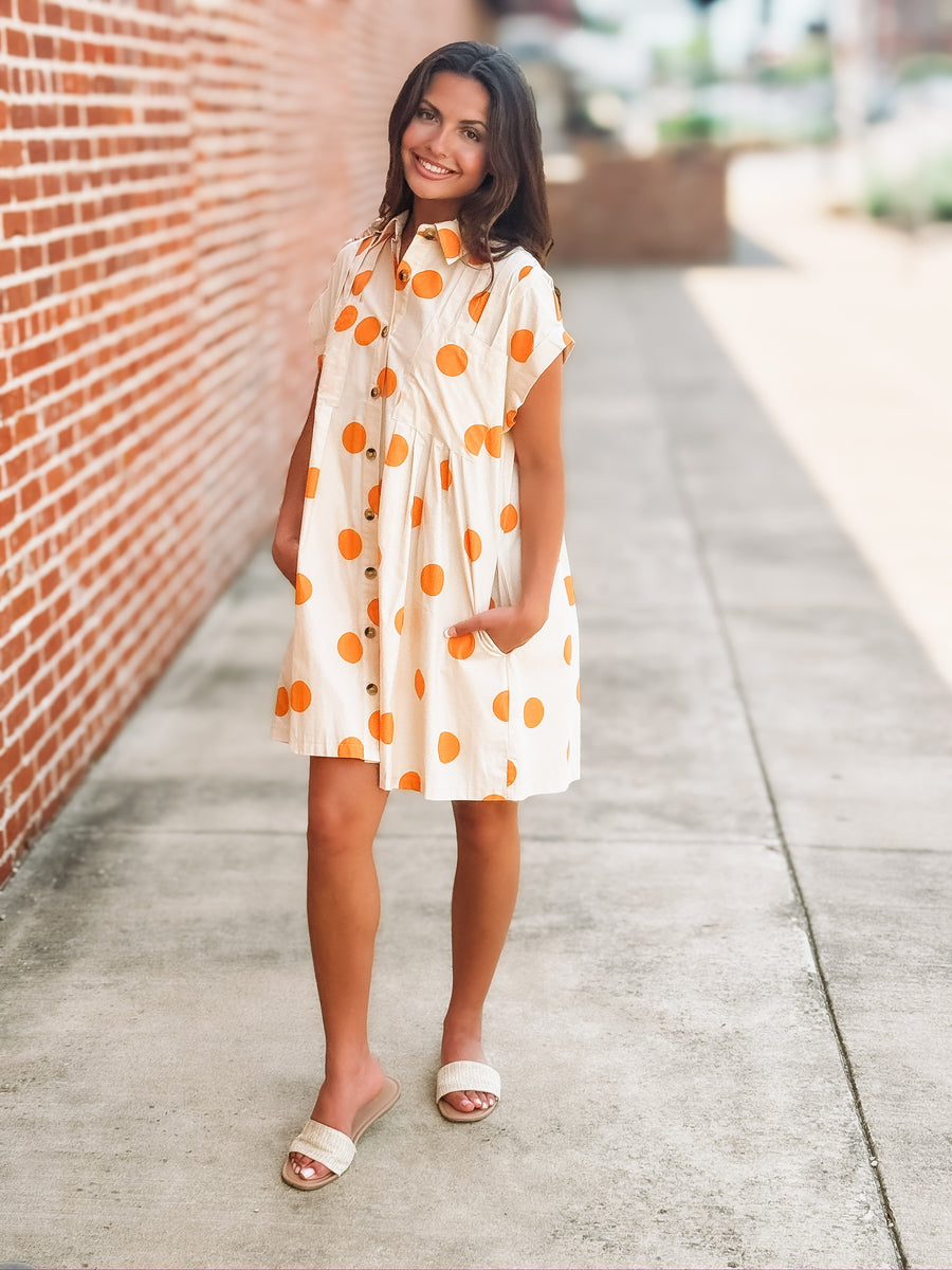 Cream mini dress with an orange polka dot print, button front closure, short sleeves, side pockets, and a relaxed fit.
