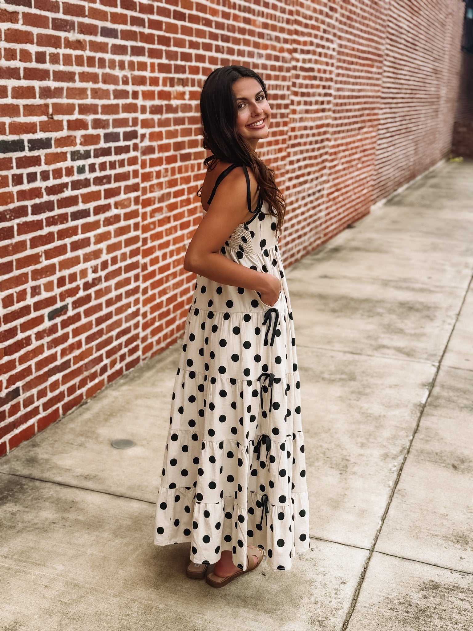Cream maxi dress with an black polka dot print, tiered skirt, fitted bust, fully smocked back, adjustable tie spaghetti straps, and bow details along the sides.