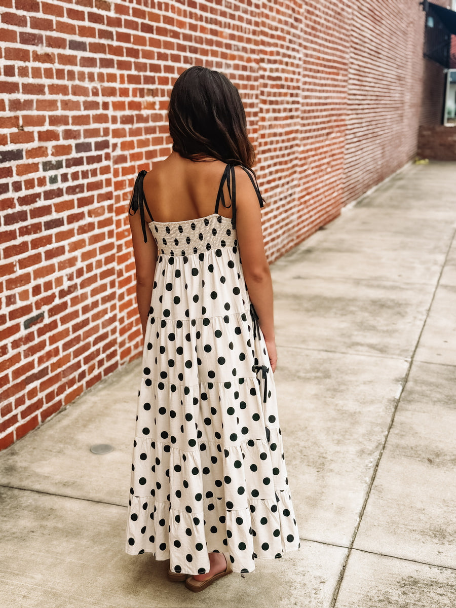 Cream maxi dress with an black polka dot print, tiered skirt, fitted bust, fully smocked back, adjustable tie spaghetti straps, and bow details along the sides.