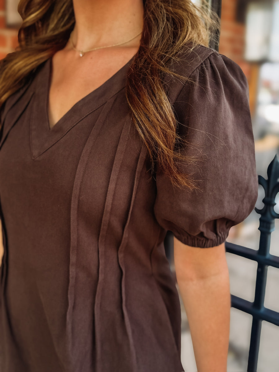 Brown denim mini dress with a v neckline, short puff sleeves, and pleated side details. 