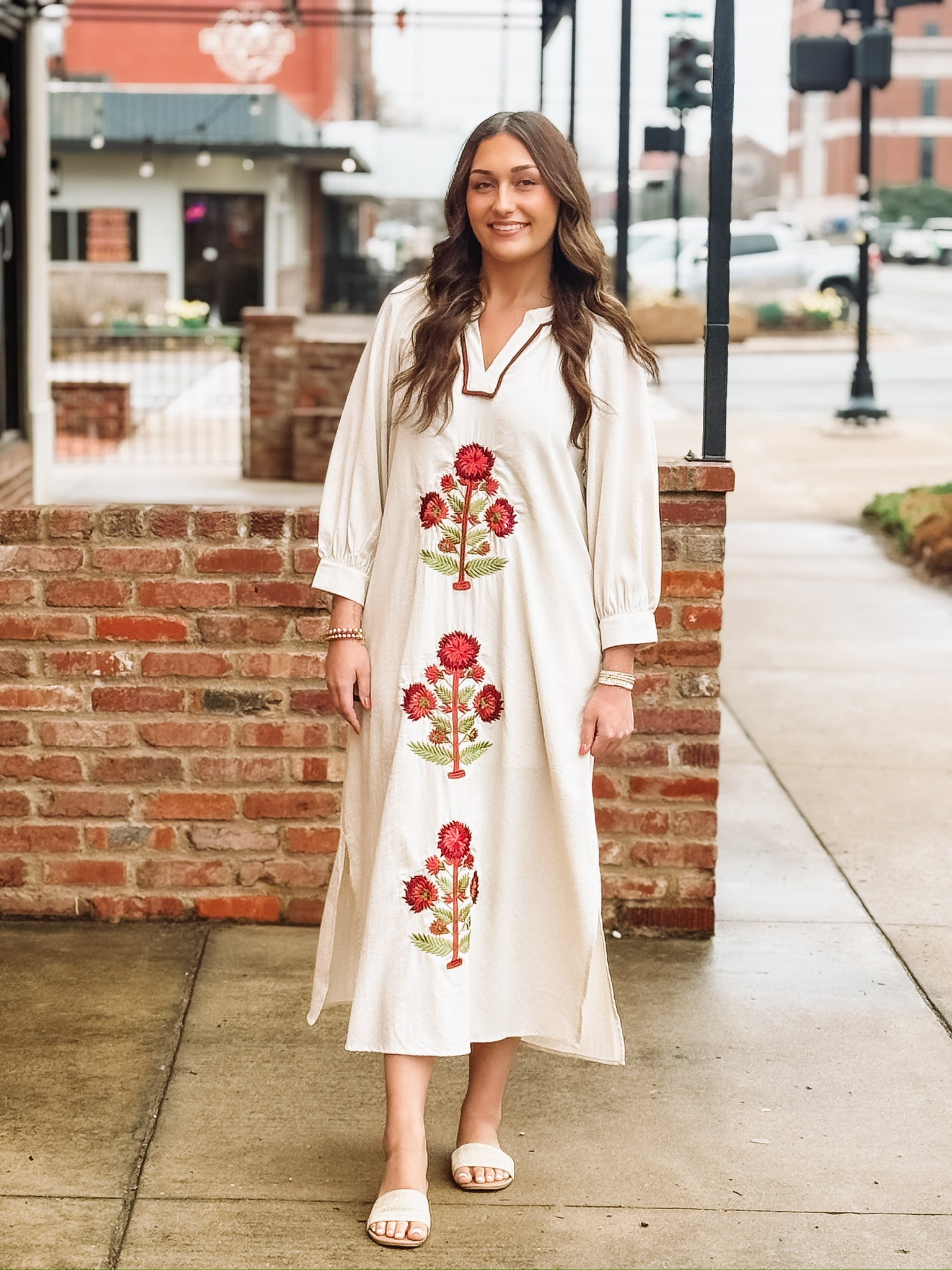 Loose, ankle length cream dress with three quarter puff sleeves and a split neckline. The dress features bold red floral embroidery with green stems arranged vertically down the front. It has a relaxed, flowy silhouette with side slits at the hem. 