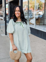 Woman wearing a green and white striped dress holding a straw bag on a sidewalk.