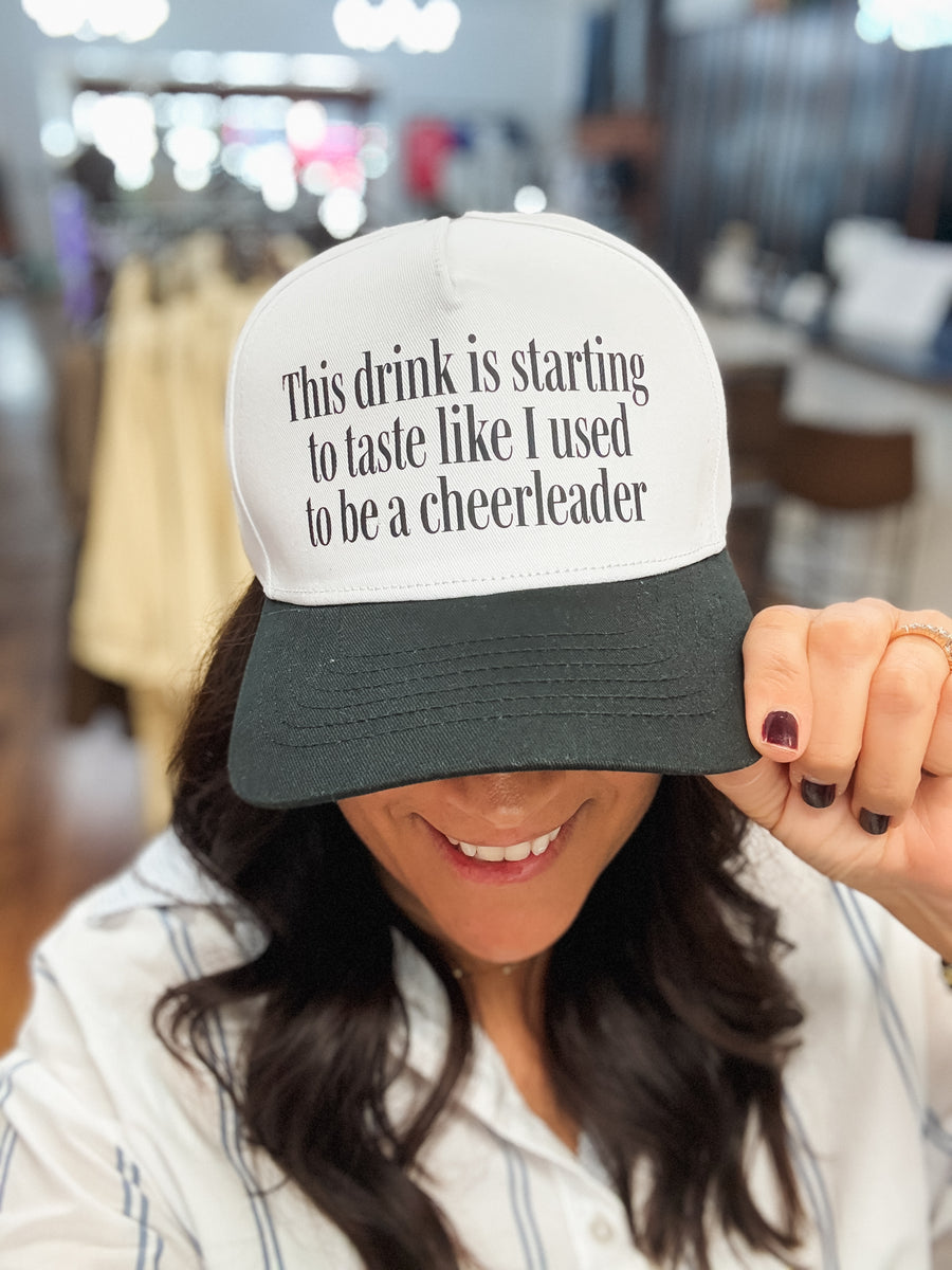 A woman wearing a white and black trucker hat with the text 'This drink is starting to taste like I used to be a cheerleader' in black lettering. She is holding the brim down with her hand in a store setting.
