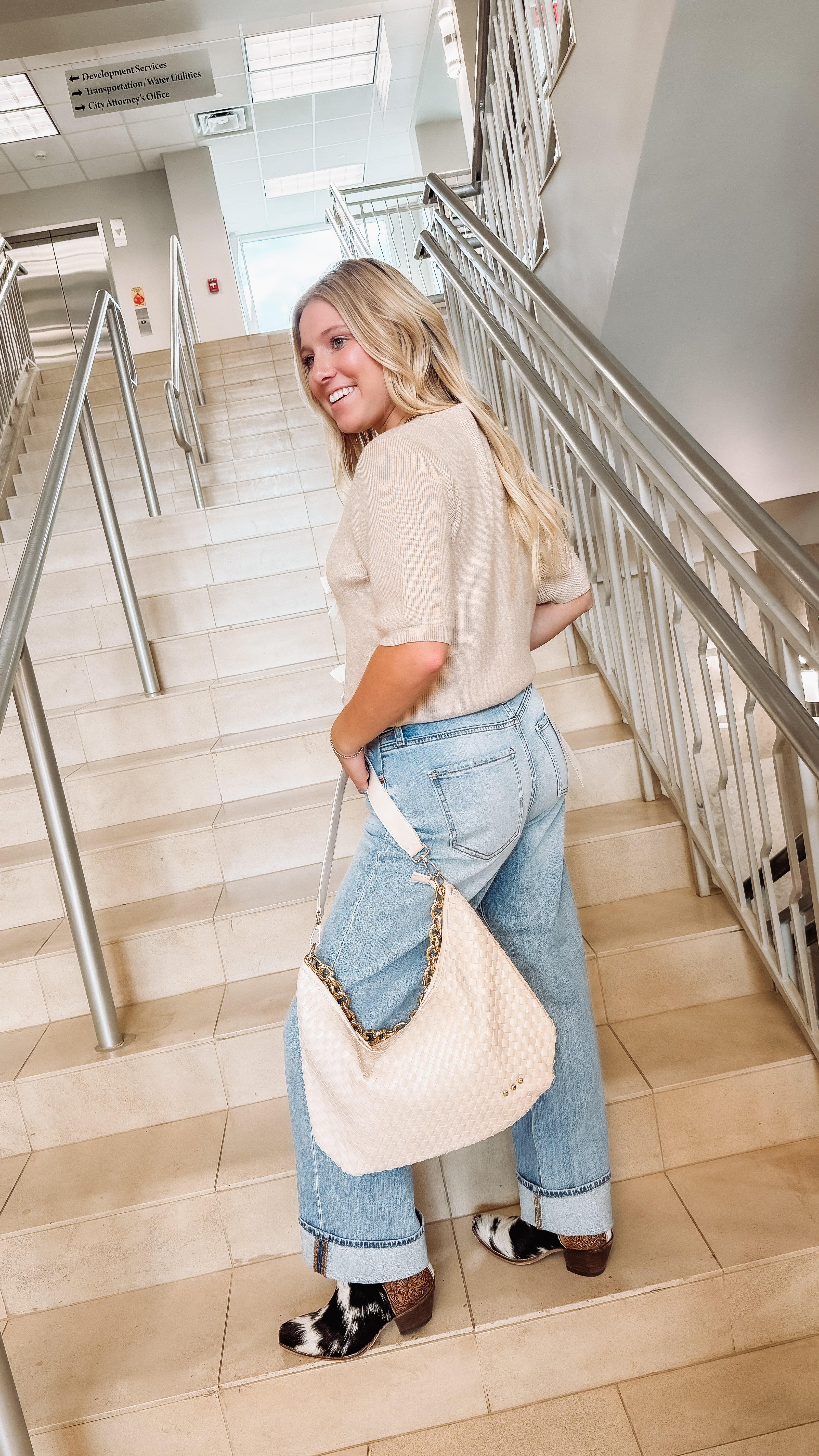 Woman walking up a staircase holding a beige handbag in a beige cardigan and jeans