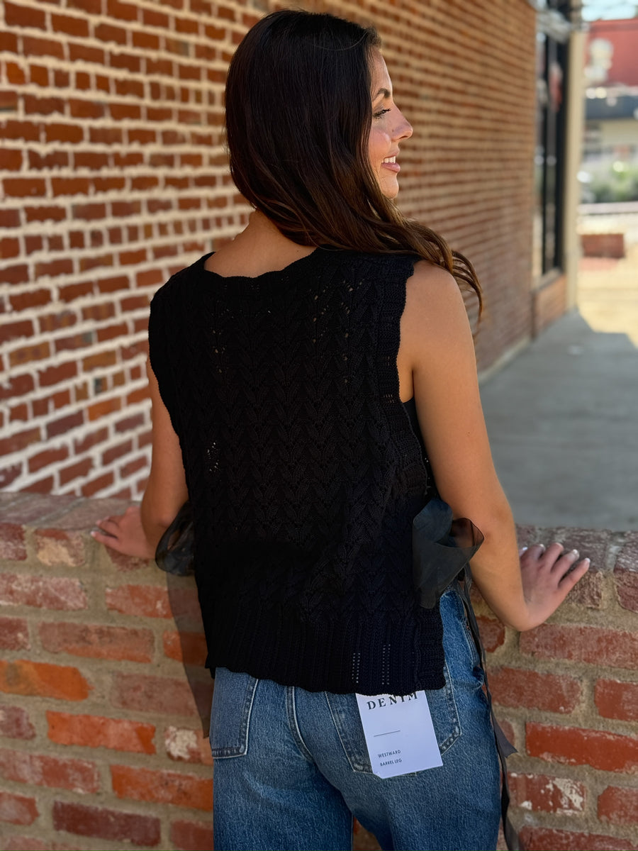 Black textured knit sweater vest with a v-neckline, ribbed trim, and open sides finished with adjustable sheer tulle tie details. Lightweight knit that can be worn alone or layered.
