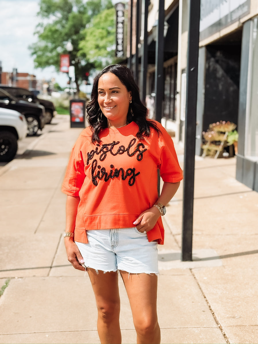 “Pistols Firing” printed in black script on an orange boxy top across the front, styled in an OSU game day theme.