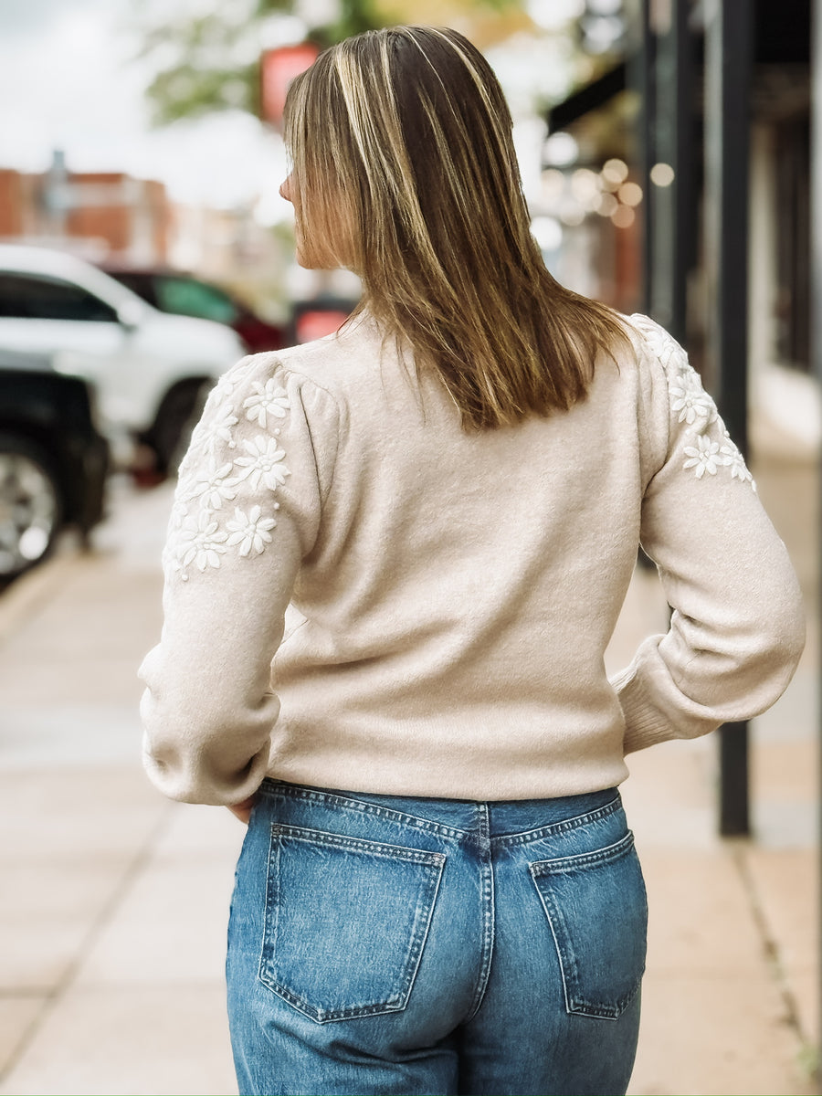 Woman wearing a beige sweater with white floral embroidered  sleeves.
