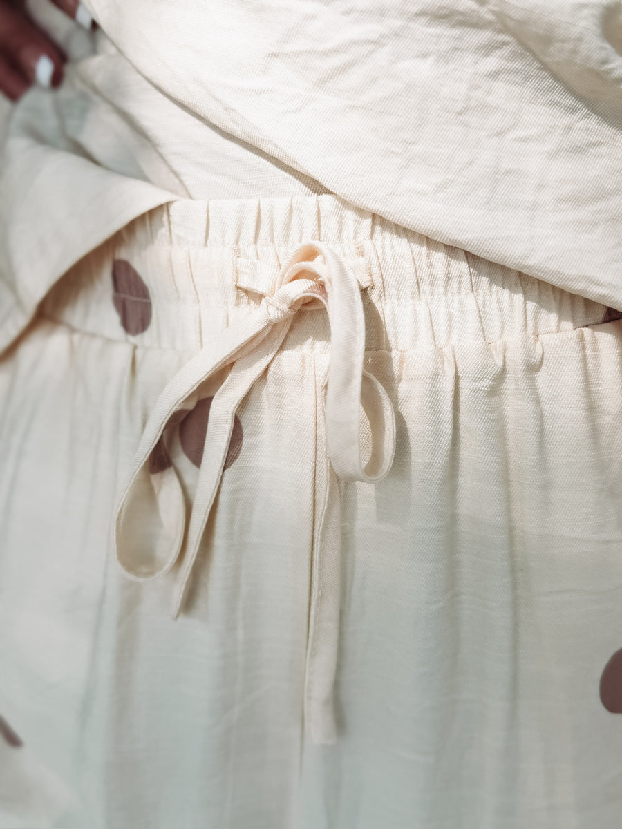 Strapless ecru matching pant  set featuring a flowy tube top with an elastic neckline and wide leg pants with an elastic drawstring waistband, pockets, and a soft light brown polka dot print.