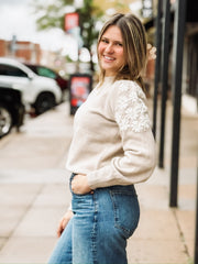 Woman wearing a beige sweater with white floral embroidered sleeves.