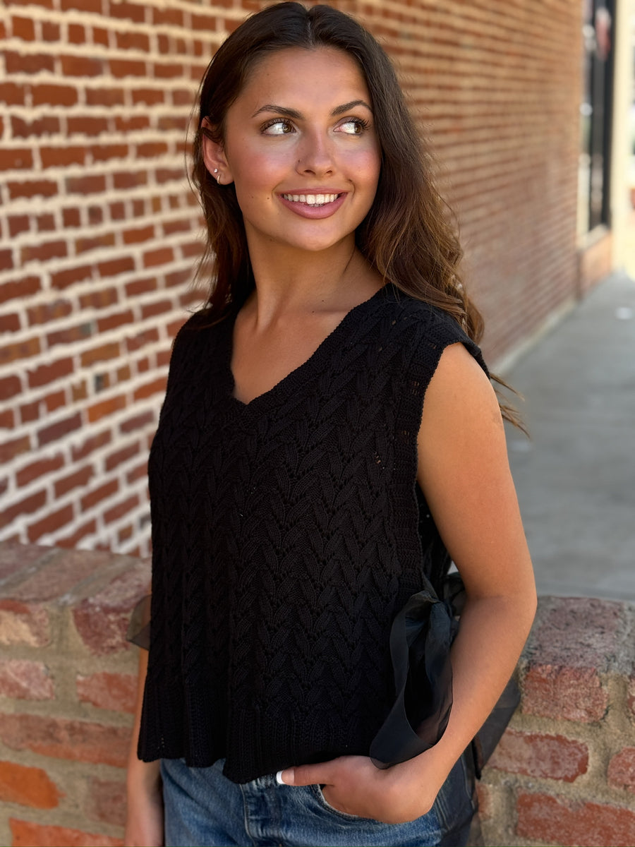 Black textured knit sweater vest with a v-neckline, ribbed trim, and open sides finished with adjustable sheer tulle tie details. Lightweight knit that can be worn alone or layered.