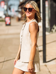 Woman wearing a beige sleeveless vest and skirt set and sunglasses on a city street.