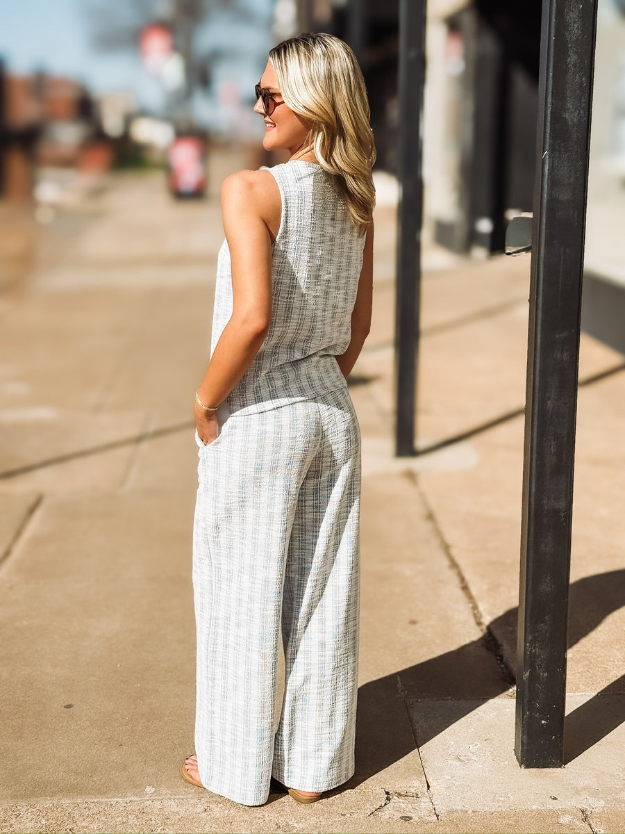 Sleeveless, textured two-piece outfit in a soft light blue and white woven fabric. The top features a round neckline, front button detailing, and a slightly relaxed, straight fit. The matching pants are high-waisted with a wide-leg silhouette
