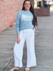 Woman wearing a blue sweater with an American flag design and white pants on a wooden deck.