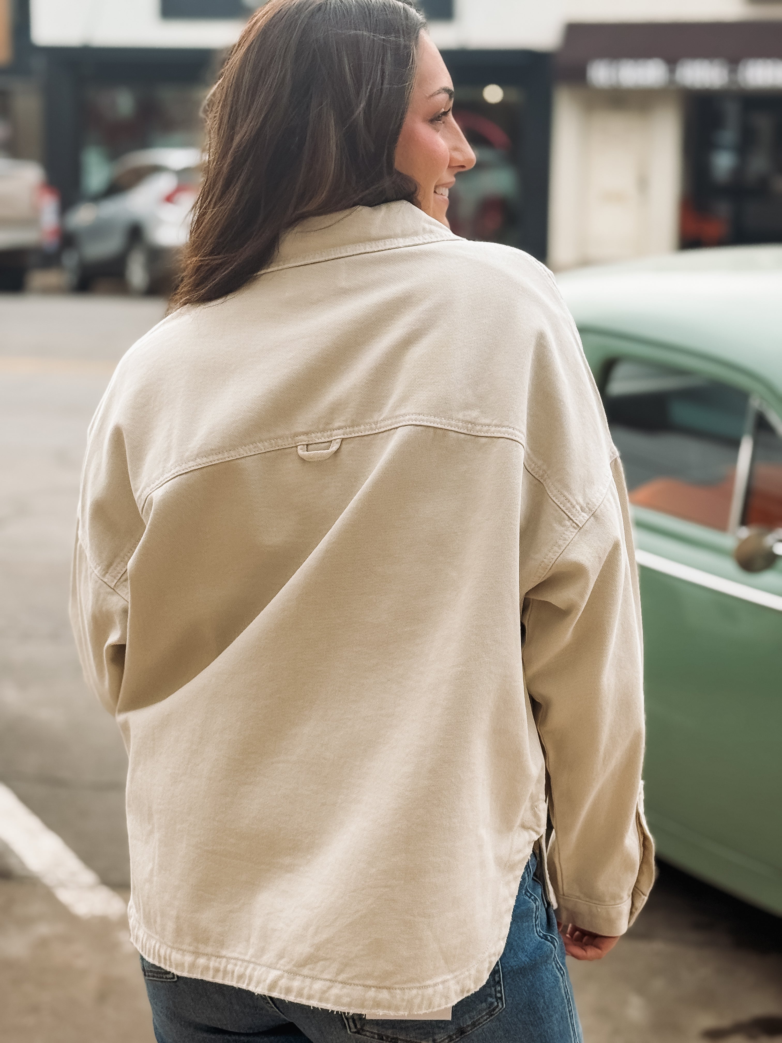 Person wearing a beige jacket standing on a street with a green car in the background