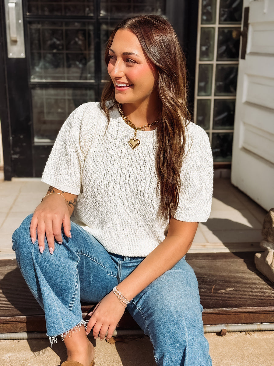 Woman sitting on a bench wearing a white top and blue jeans, smiling.