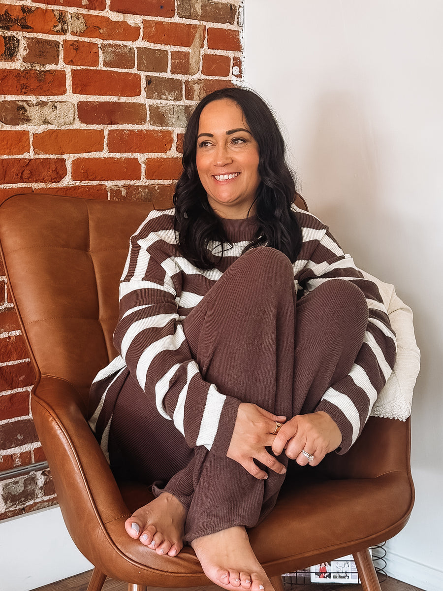 Woman sitting on a brown leather chair wearing a brown and white striped sweater and pants against a brick wall.