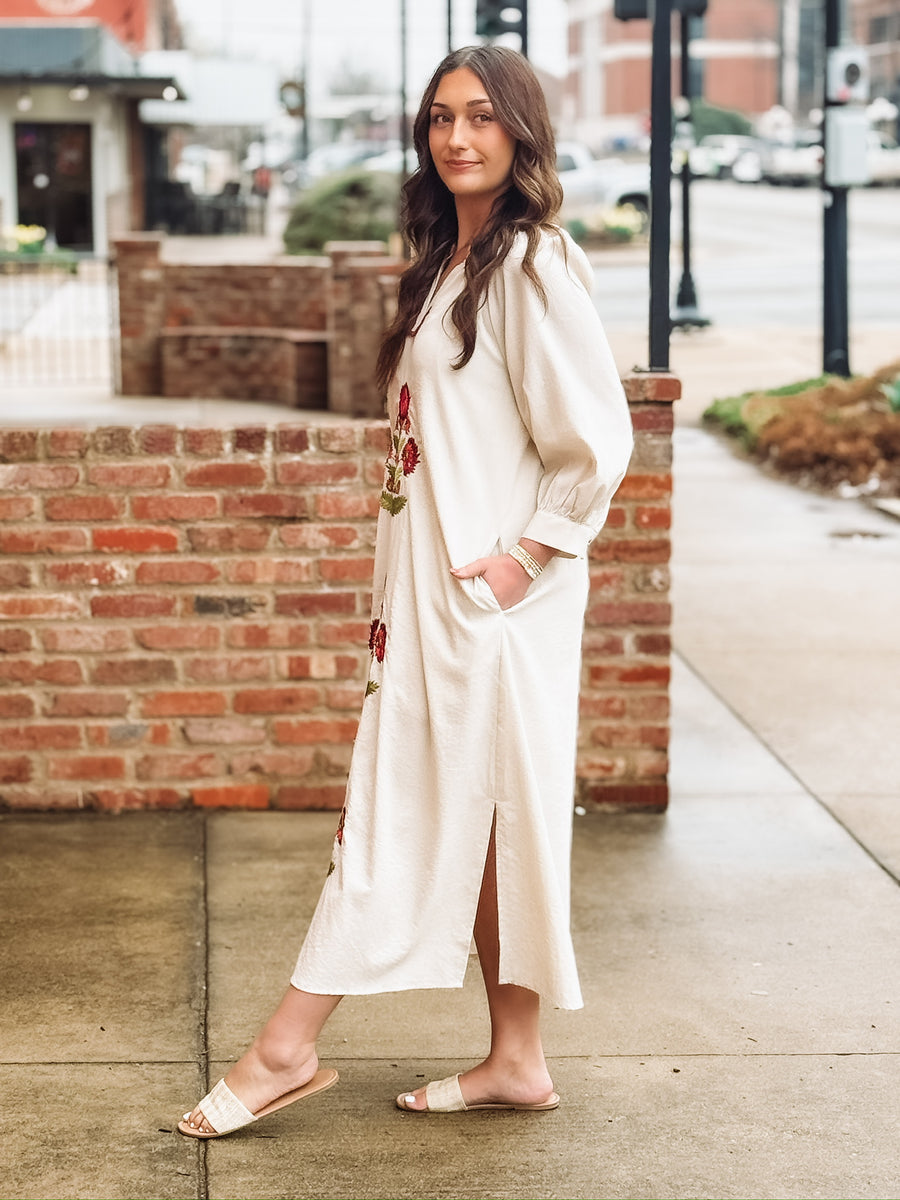 Loose, ankle length cream dress with three quarter puff sleeves and a split neckline. The dress features bold red floral embroidery with green stems arranged vertically down the front. It has a relaxed, flowy silhouette with side slits at the hem. 