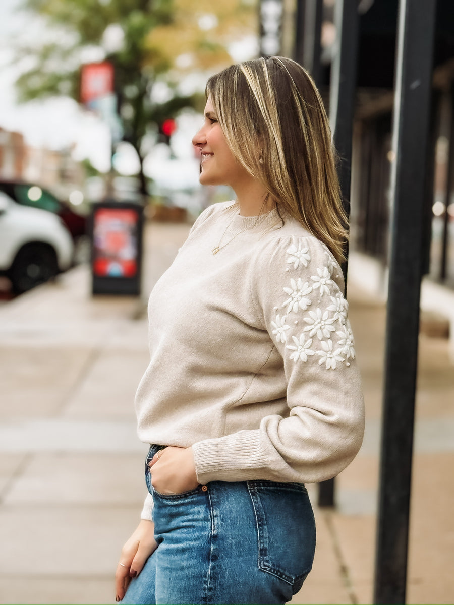 Woman wearing a beige sweater with white floral embroidered  sleeves.