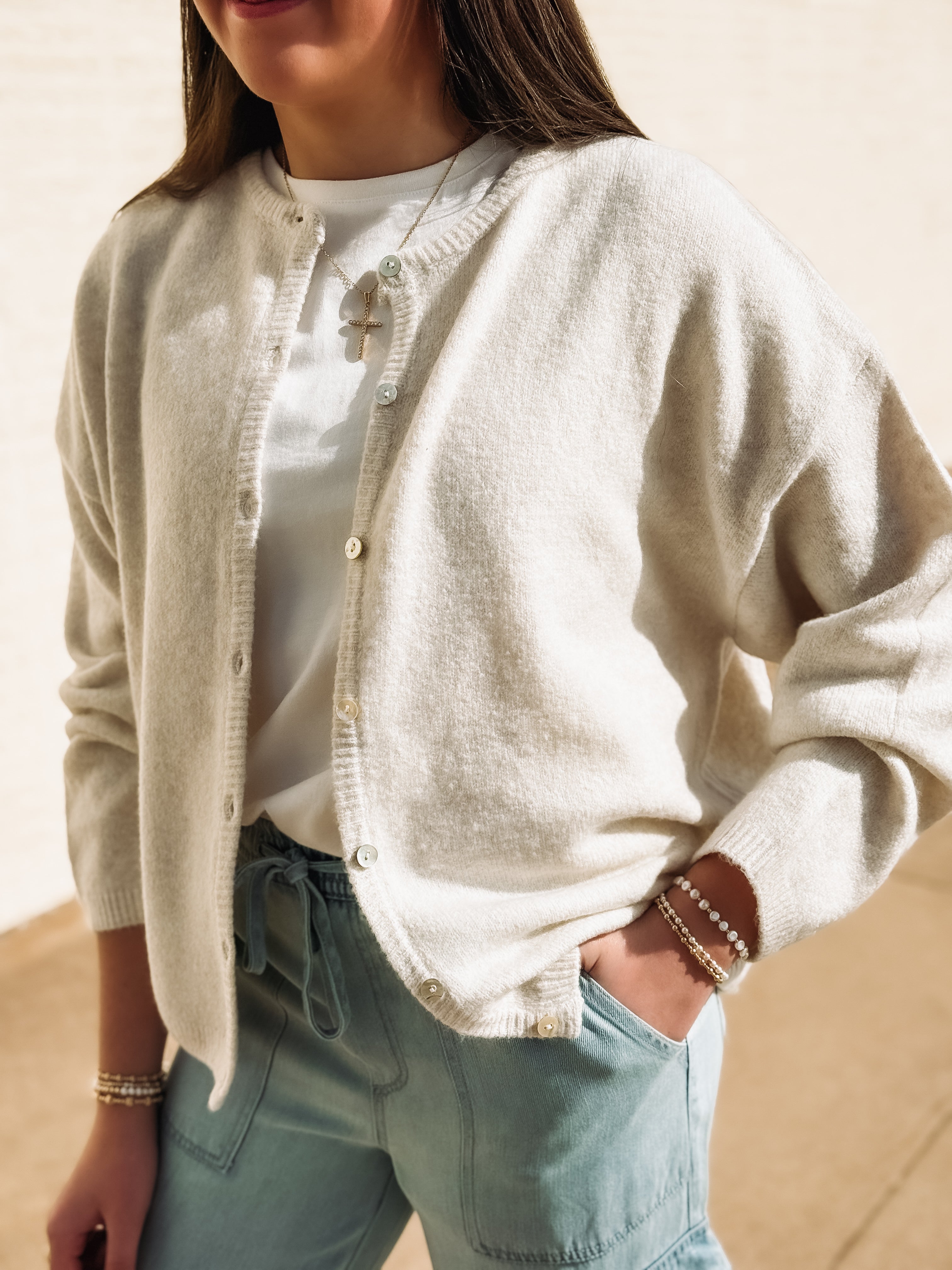 Person wearing a beige cardigan over a white shirt with light blue jeans, standing against a neutral background.
