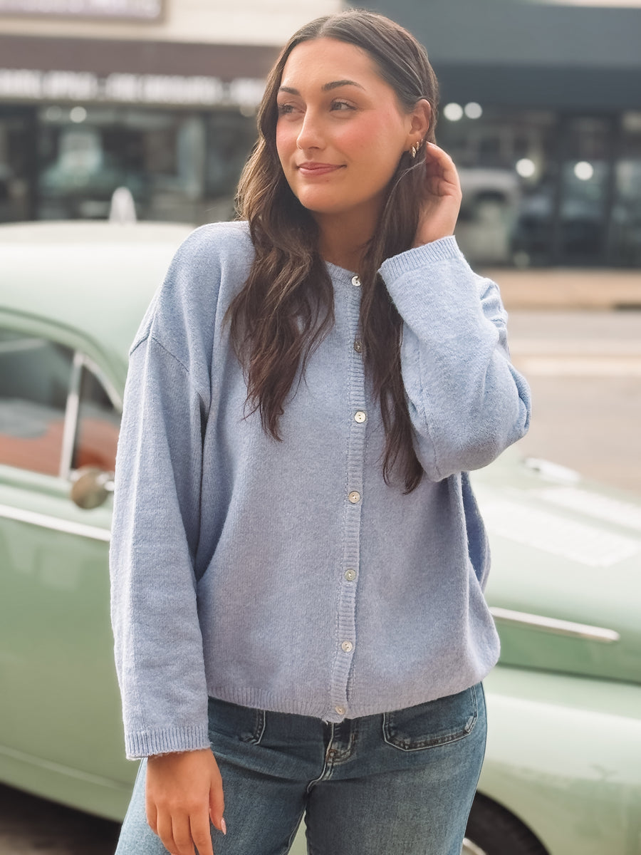 Woman wearing a light blue cardigan and jeans standing in front of a vintage car.