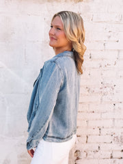 Woman wearing a light denim colored jacket standing against a light brick wall.
