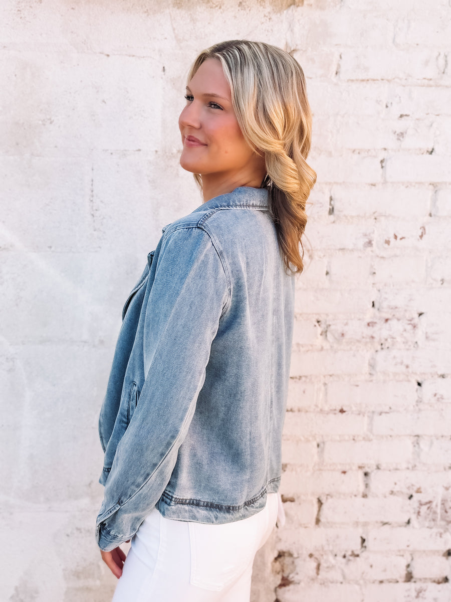 Woman wearing a light denim colored jacket standing against a light brick wall.