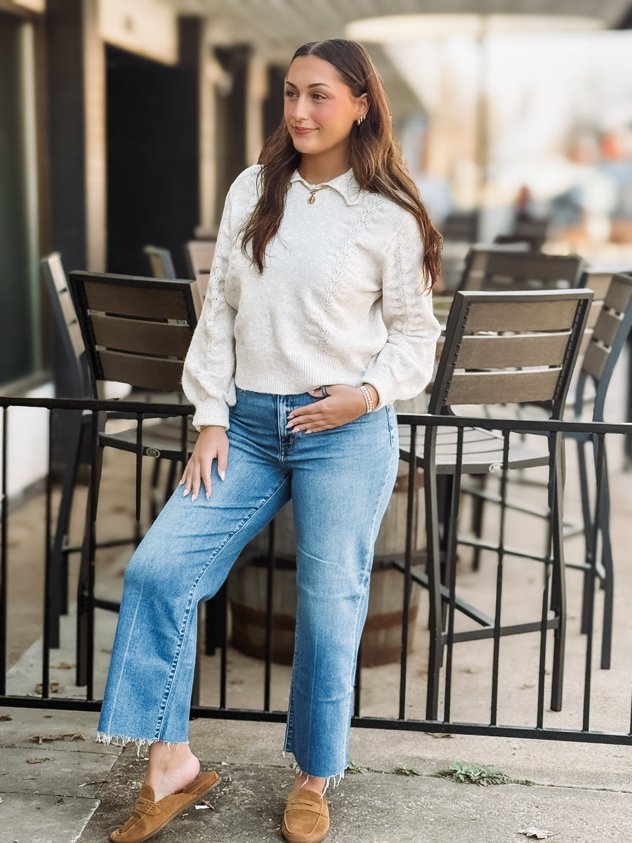 Woman wearing a cream sweater and blue jeans standing outdoors.