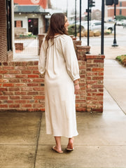 Loose, ankle length cream dress with three quarter puff sleeves and a split neckline. The dress features bold red floral embroidery with green stems arranged vertically down the front. It has a relaxed, flowy silhouette with side slits at the hem. 