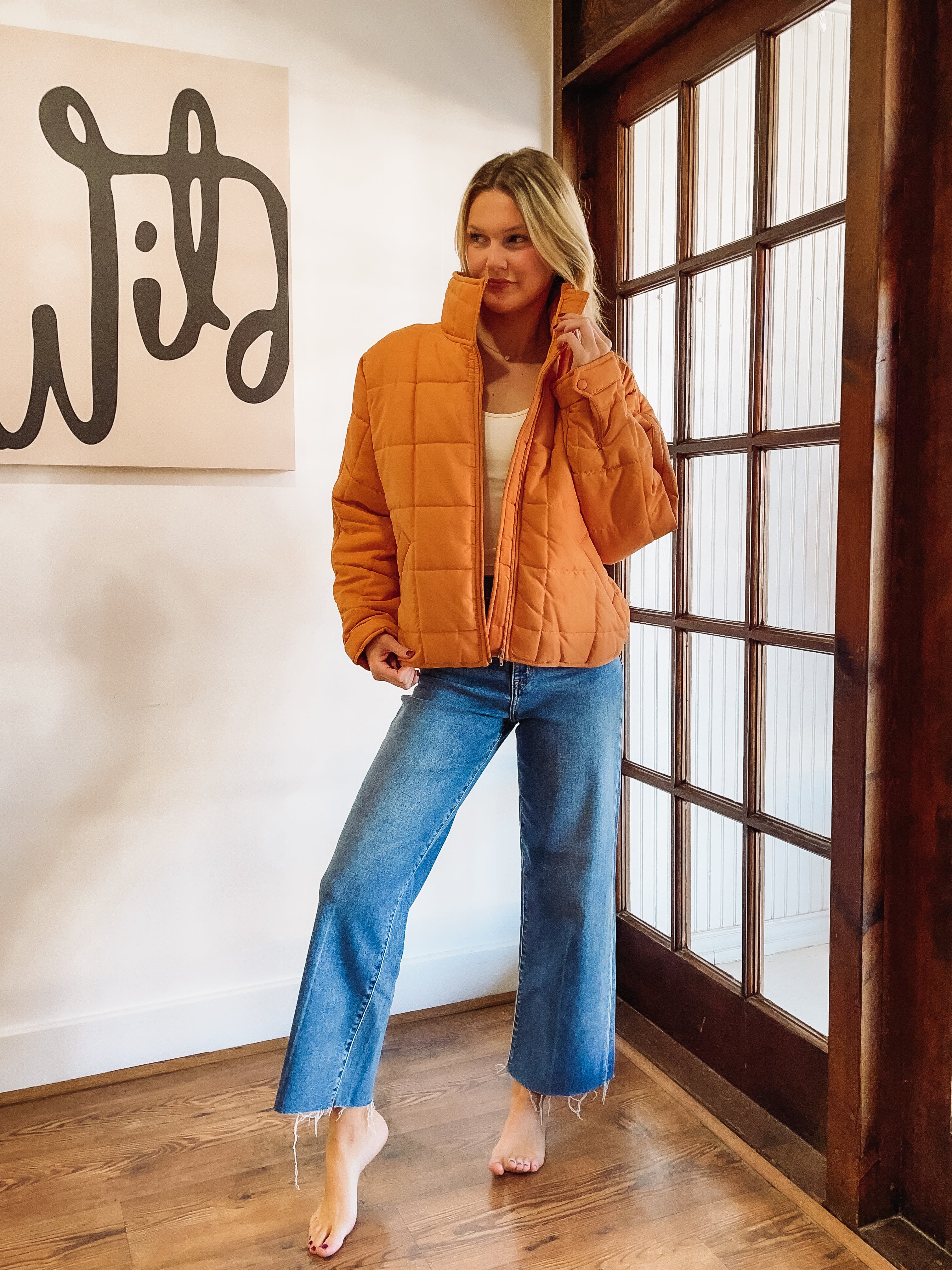Woman wearing an orange puffer jacket and blue jeans indoors.