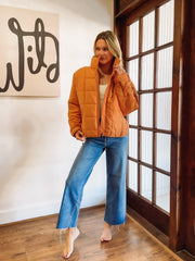 Woman wearing an orange puffer jacket and blue jeans indoors.