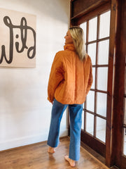 Person wearing an orange jacket and blue jeans standing in a room with wooden flooring and a large window.