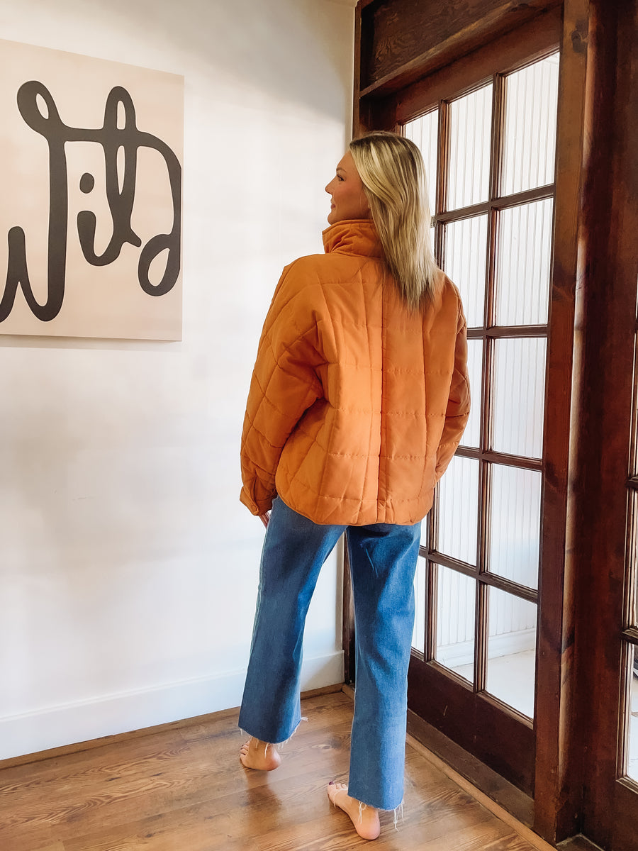 Person wearing an orange jacket and blue jeans standing in a room with wooden flooring and a large window.