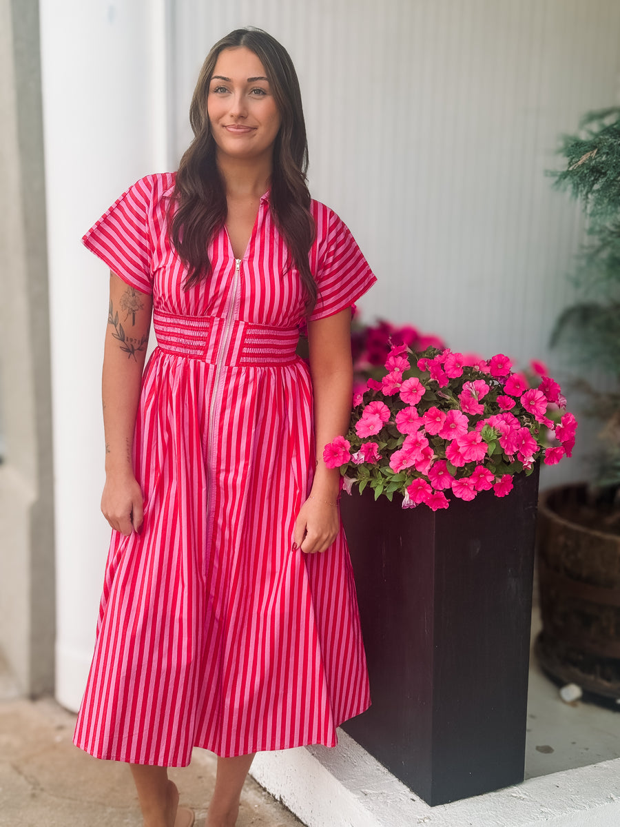 Woman in a pink and red striped dress standing next to a potted plant with pink flowers.