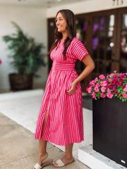 Woman in a pink and red striped dress standing next to a potted plant with pink flowers.