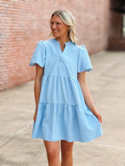 Woman wearing a light blue dress standing against a brick wall.