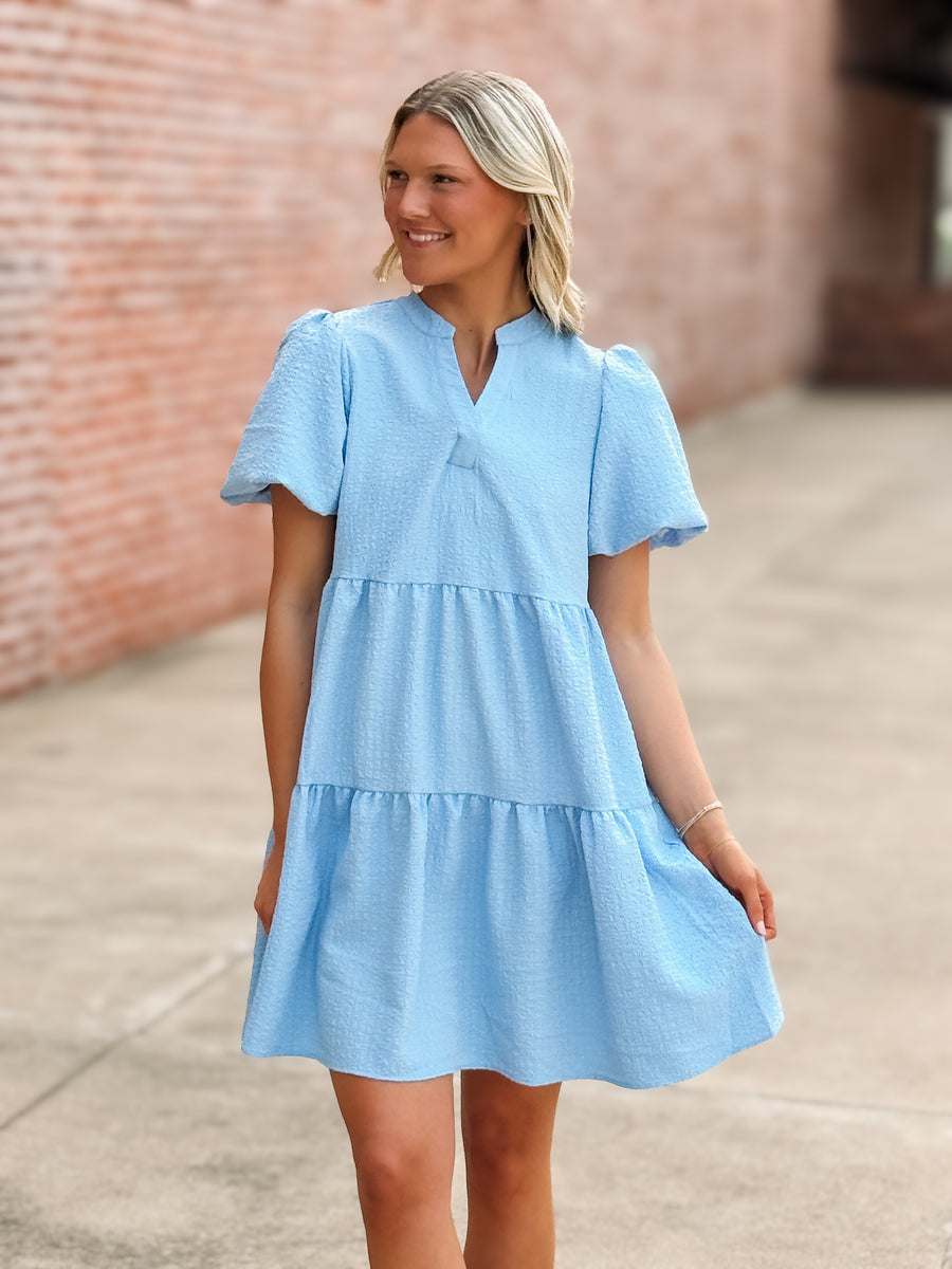 Woman wearing a light blue dress standing against a brick wall.