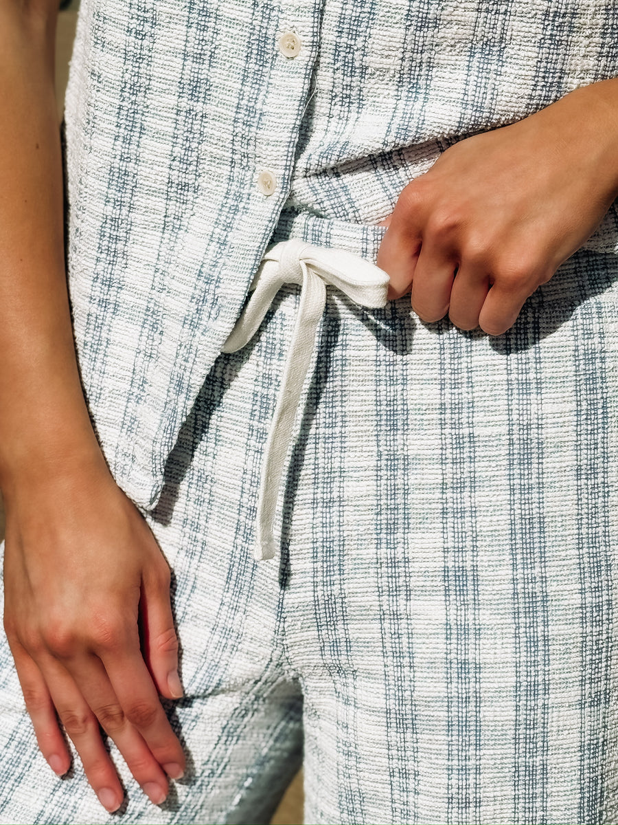 Sleeveless, textured two-piece outfit in a soft light blue and white woven fabric. The top features a round neckline, front button detailing, and a slightly relaxed, straight fit. The matching pants are high-waisted with a wide-leg silhouette