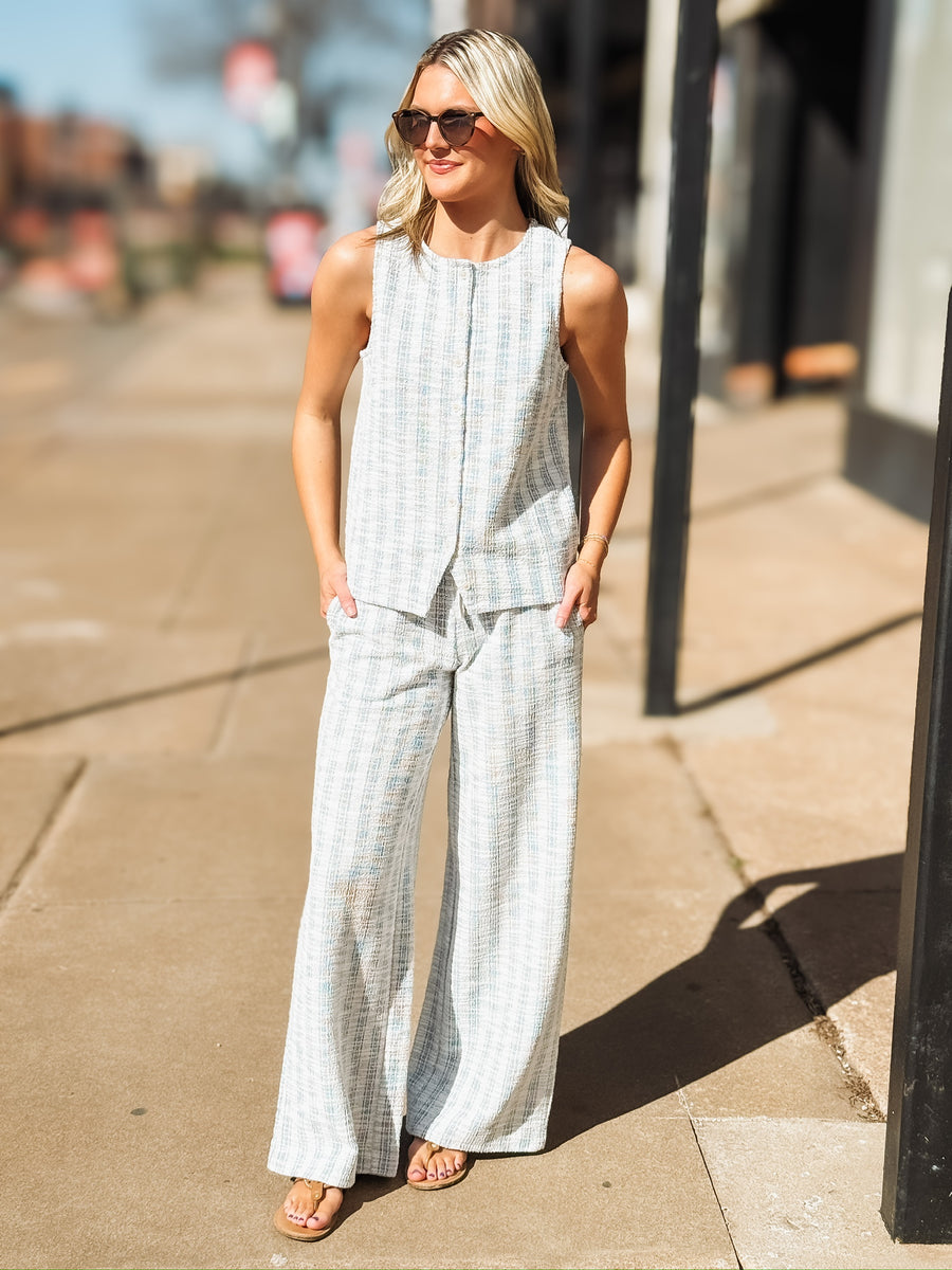 Sleeveless, textured two-piece outfit in a soft light blue and white woven fabric. The top features a round neckline, front button detailing, and a slightly relaxed, straight fit. The matching pants are high waisted with a wide leg cut.