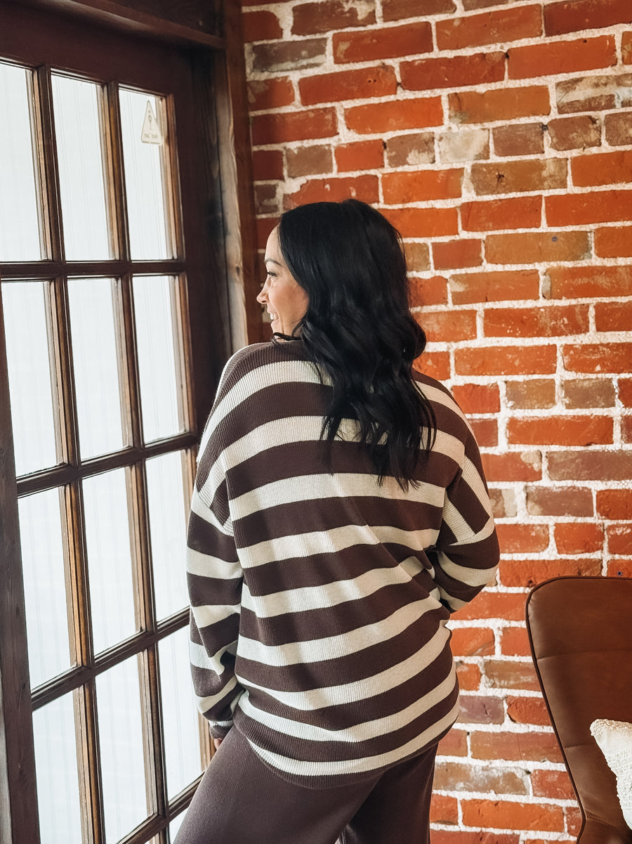 Woman standing in a room with a brick wall and large window in a chocolate and cream striped lounge top with matching brown pants.